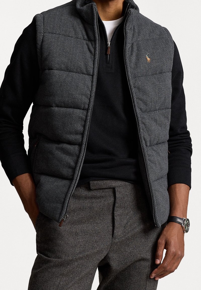 Gilet imbottito grigio a motivo a zigzag, con colletto nero con zip, indossato sopra una maglietta nera a maniche lunghe e pantaloni grigi, completo di un orologio.