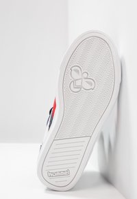 Suela de zapatilla blanca con textura de espiga, que presenta un logo en el centro y acentos de color rojo y azul en la parte superior.