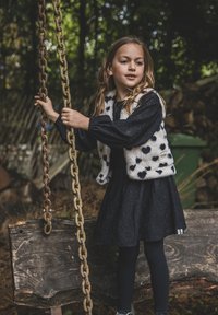 Jong meisje in zwarte jurk en wit gespikkelde vest die roestige schommelkettingen vasthoudt in een bosrijke buitenomgeving.
