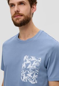 Blaues Baumwoll-T-Shirt mit kurzen Ärmeln, das ein weißes Blattmuster im Taschenbereich, einen runden Halsausschnitt und eine glatte Textur aufweist.