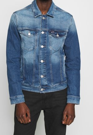 Mężczyzna w średnio niebieskiej kurtce dżinsowej na guziki z dwoma kieszeniami na piersi, ubrany w czarne spodnie, na jednolitym tle.