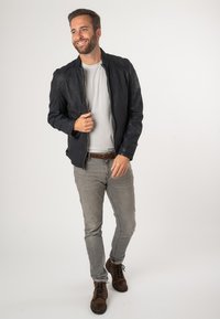 Giacca di pelle nera con dettagli testurizzati sulle spalle, indossata sopra una t-shirt grigia chiara. Un paio di jeans grigi e stivali marroni con lacci sono visibili.