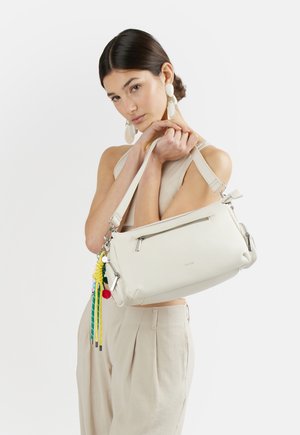 Giovane donna con capelli scuri raccolti in uno chignon, indossa abiti beige e grandi orecchini, tiene una borsa bianca con nappe colorate.
