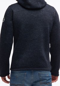 Granatowa bluza z kapturem o fakturowanym wzorze, z kapturem, detalem bocznego zamka i ściągaczami przy mankietach. Noszona na jasnoniebieskie dżinsy.