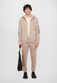 Sudadera beige con cremallera y mangas texturizadas, combinada con pantalones cargo a juego. La modelo sostiene un bolso tote negro y lleva zapatillas negras.