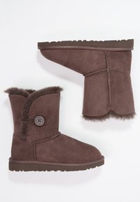 Brązowe zamszowe botki za kostkę z miękką podszewką z sztucznego futra, akcentem guzika oraz chropowatą gumową podeszwą. Posiadają falbankę na górnej krawędzi.