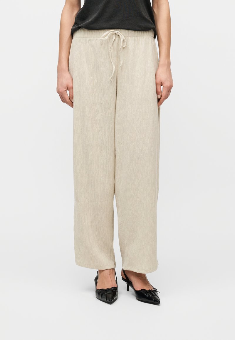 Μπεζ παντελόνι φαρδιάς γραμμής με κορδόνι στη μέση, συνδυασμένο με μαύρες γόβες slingback με μυτερή μύτη και μαύρο τοπ, φαίνεται από τη μέση έως τα πόδια.