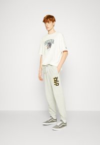 Hvid t-shirt med grafisk design, lysegrå sweatpants med sorte og gule accenter, og grå sneakers. Model står på en neutral baggrund.