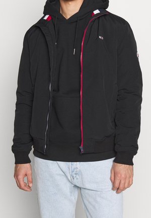 Homme portant un sweat à capuche noir et une veste noire zippée avec des accents rouges et blancs, assortis à un jean bleu clair.