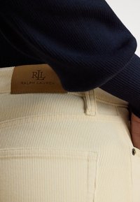 Jasnobeżowe spodnie sztruksowe z pionowymi żebrowaniami, z skórzaną metką z tłoczonym napisem "RLL RALPH LAUREN" na pasku. Powyżej widoczny ciemny sweter.