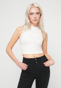 Ung kvinne med langt blondt hår iført en ermeløs hvit crop top og høy midje svarte jeans, med hendene i lommene, nøytral bakgrunn.