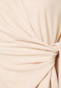 Tecido bege com uma textura suave, apresentando um design torcido na cintura que cria pregas drapeadas. Detalhe de costura subtil visível.