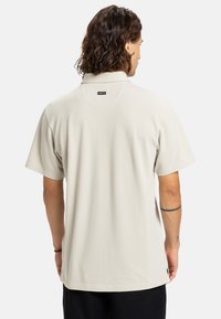 Kortærmet polo i lys beige stof med blød tekstur, der har en klassisk krave og et mærke nær halsudskæringen.
