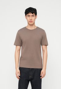 Νέος άνδρας με κοντά μαύρα μαλλιά που φοράει ένα απλό καφέ κοντομάνικο μπλουζάκι και σκουρόχρωμο τζιν, στέκεται μπροστά σε λευκό φόντο.