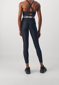 Kobieta w czarnym krzyżowym staniku sportowym, czarnych legginsach o wysokim stanie z logo w pasie oraz czarnych butach sportowych z akcentami w odcieniach turkusu i fioletu.