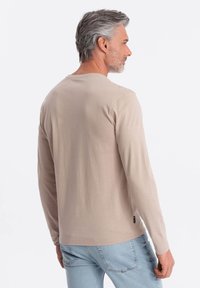 Långärmad beige t-shirt i mjuk bomull, med rundad halsringning och en figursydd design, kombinerad med ljusblå jeans.