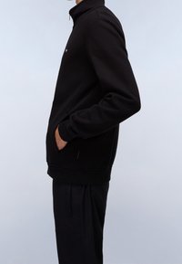 Schwarze Zip-Jacke mit hohem Kragen, ausgestattet mit gerippten Bündchen und Saum, seitlichen Taschen und einem kleinen Logo auf der Brust. Weicher Stoff. 