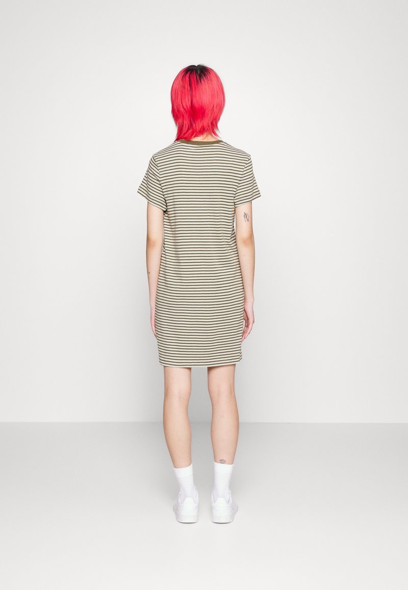 Personne aux cheveux rouge vif portant une robe beige courte à manches courtes, longueur genou, avec des rayures horizontales noires, des chaussettes blanches et des chaussures blanches, vue de dos.