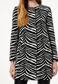 Mujer vestida con un abrigo largo a rayas de cebra en blanco y negro, con mangas largas, de pie con los brazos relajados a los lados.