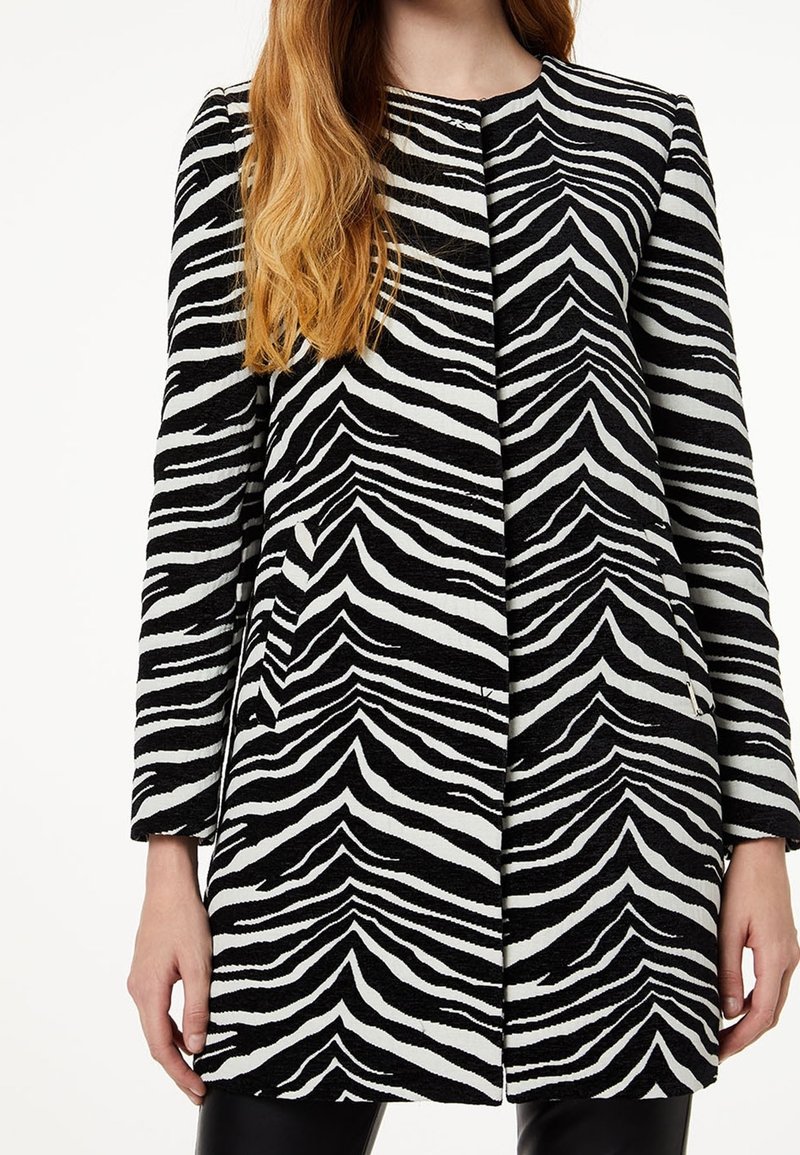 Mujer vestida con un abrigo largo a rayas de cebra en blanco y negro, con mangas largas, de pie con los brazos relajados a los lados.