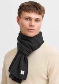 Sciarpa a maglia grigia scura con una texture a coste, con un piccolo patch con il marchio, appoggiata attorno al collo di una persona in un maglione beige.
