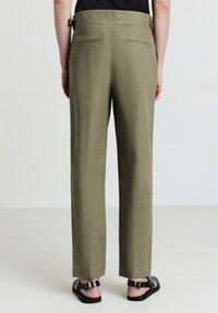 Pantaloni verde oliva a vita alta con gamba ampia, caratterizzati da una texture liscia, tasche posteriori e passanti per cintura, abbinati a sandali scuri con cinturini.