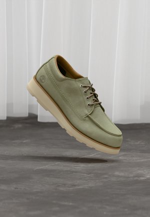 Vihreä mokkanahkainen kenkä, jossa on sileä pinta, pyöreä kärki ja ruskeat nauhat. Kenkässä on paksu kermaan vivahtava pohja ja logo sivulla.