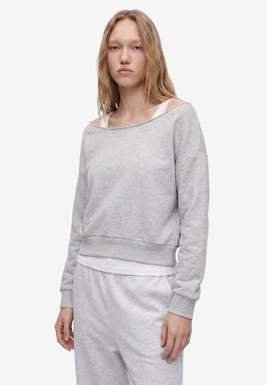 Ung kvinde med langt blondt hår iført lysegrå off-shoulder sweatshirt og matchende sweatpants mod en hvid baggrund.