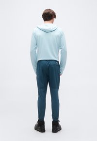 Άνδρας που στέκεται με την πλάτη γυρισμένη, φορώντας ανοιχτό μπλε φούτερ με κουκούλα, τιρκουάζ αθλητικό παντελόνι και μαύρα ορειβατικά μποτάκια σε λευκό φόντο.