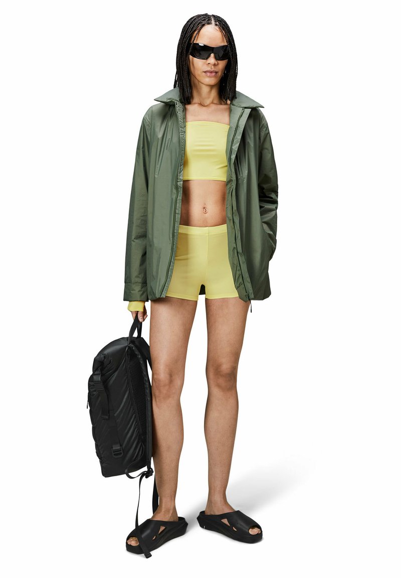 Un mannequin porte une veste légère verte, un crop top jaune et un short, tenant un sac à dos noir, associé à des chaussures noires à enfiler.