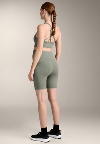 Olive zelena športna nedrček s križnimi naramnicami, v kombinaciji z usklajenimi visokimi kolesarskimi šortsami. Črne športne čevlje zaokrožajo outfita.