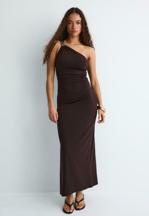 Kobieta z długimi kręconymi włosami w dopasowanej, asymetrycznej, ciemnobrązowej sukni maxi i czarnych sandałach, stojąca na jasnym, jednolitym tle.