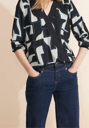 Mujer con blusa de manga arremangada con patrón geométrico en blanco y negro y jeans de mezclilla azul oscuro, con las manos en los bolsillos.