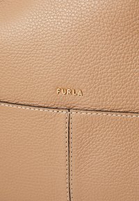 Bež trgovinska torba od kože s teksturiranom površinom, s pozlaćenim urezanim logom "FURLA" i kontrastnim bijelim šavom duž šava.