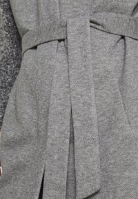 Tessuto grigio con una finitura morbida e testurizzata, caratterizzato da un design avvolgente e una cintura legata in vita. Linee pulite e dettagli minimali.
