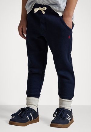 Kind draagt marineblauwe joggingbroek met een witte trekkoord, witte sokken en marineblauwe sneakers met bruine zolen, staat met handen in de zakken.