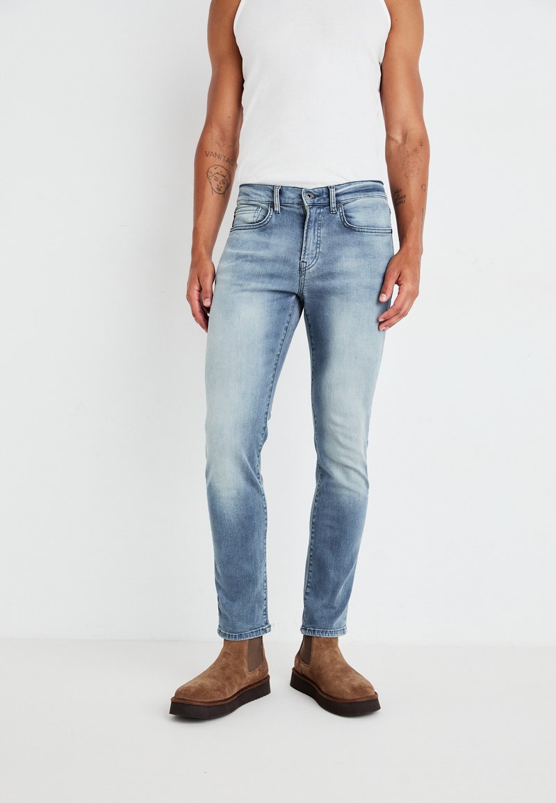 Jeans de denim azul claro con un ajuste slim, que cuentan con bolsillos frontales y un acabado desgastado; combinados con botas de tobillo de ante marrón.