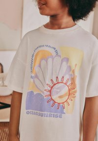Hellbeige T-Shirt mit einem bunten Grafikdesign eines Sonnenaufgangs über dem Ozean, das Muscheln und Korallen zeigt. Mit bestickten Sonnendetails und fettem Text am unteren Rand.
