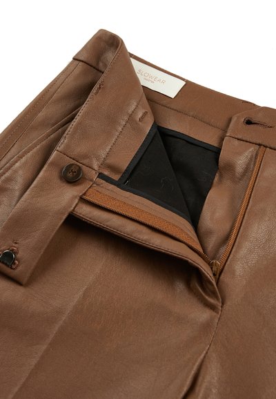 Bruine leren broeken met een gladde textuur, voorzien van een ritszak, knoopsluiting en zwarte binnenvoering. Merkenlabel zichtbaar.