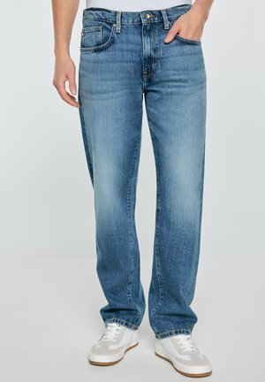 Mężczyzna noszący luźne niebieskie dżinsy i białe tenisówki, stoi z jedną ręką w kieszeni na jednolitym tle.