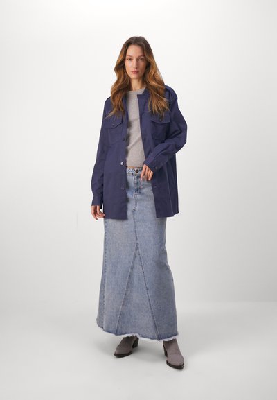 Marineblauwe oversized jas met twee borstzakken, gecombineerd met een lichtgrijze trui en een lange spijkerrok met een voornaad. Grijze enkelbotten.