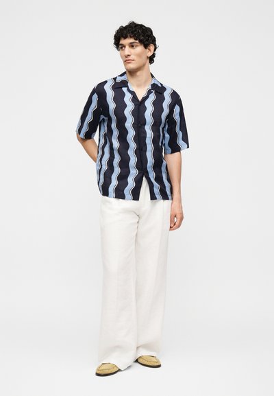 Άνδρας με σγουρά μαλλιά που φοράει κοντομάνικο μαύρο και μπλε πουκαμισάκι με κυματιστές ρίγες, λευκό παντελόνι με φαρδιά μπατζάκια και μπεζ παπούτσια, στέκεται μπροστά σε λευκό φόντο.