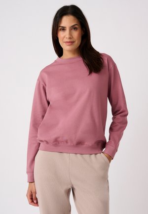 Donna con capelli scuri che indossa una felpa mauve a maniche lunghe e pantaloni beige chiaro, in piedi con una mano in tasca contro uno sfondo chiaro.