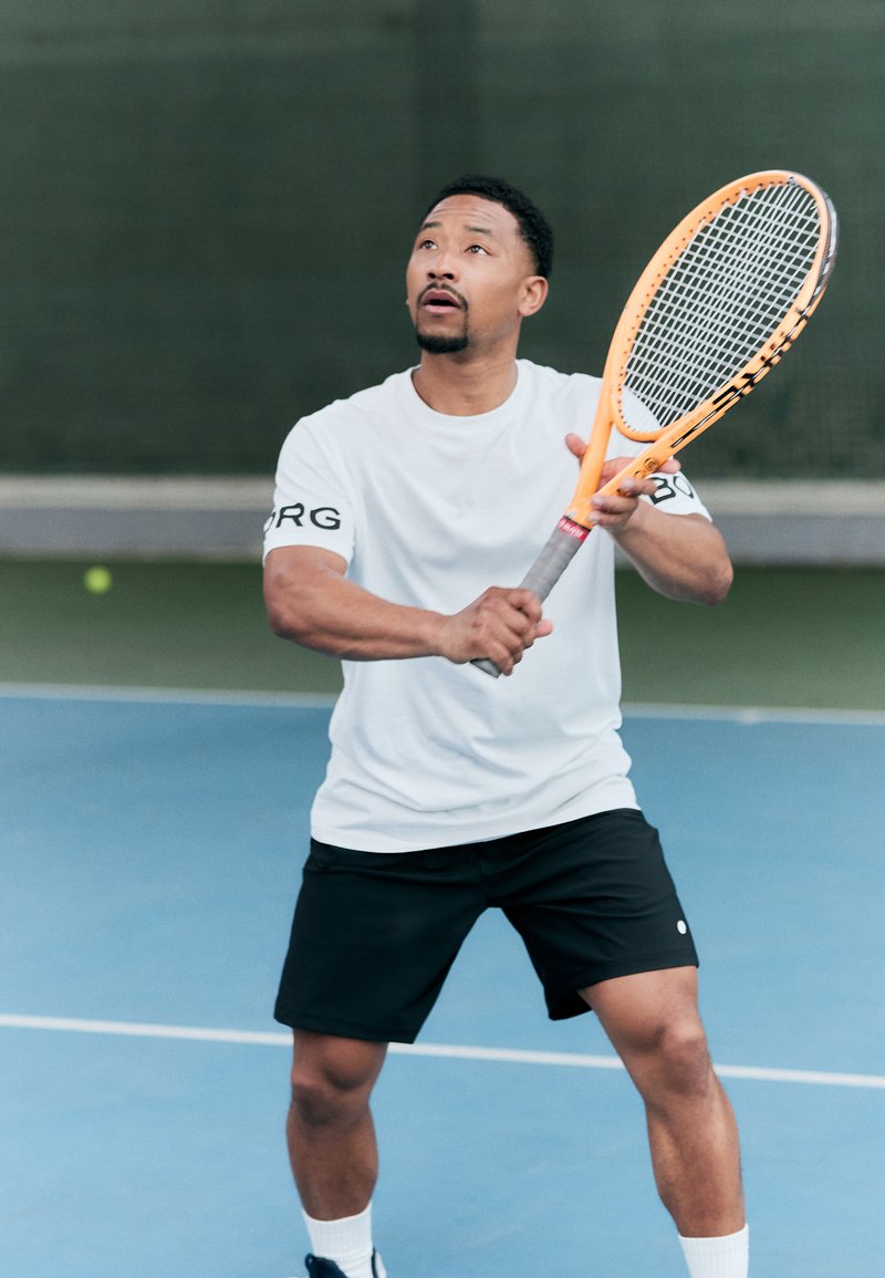 Tennisspiller i hvit skjorte med svarte detaljer og svarte shorts, holder en oransje tenniskølle, på en blå bane med grønn nett.