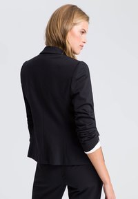 Black tailored blazer with structured shoulders, slim fit, and rolled-up sleeves revealing white cuffs. Smooth fabric, minimalistic design.