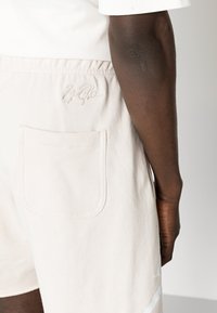 Pantaloni corti in cotone beige con una firma ricamata, dotati di una tasca posteriore e una texture morbida. Design pulito con una vestibilità rilassata.