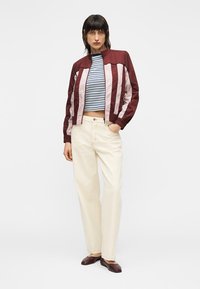 Bordeaux bomberjakke med lyserøde striber, kombineret med en striped crop top og cremefarvede bukser. Modellen har brune loafers på.