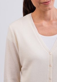 Krémová kardigán s V-výstřihem, měkkou texturou a pěti knoflíky vpředu, oblečený přes bílou vnitřní blůzu.