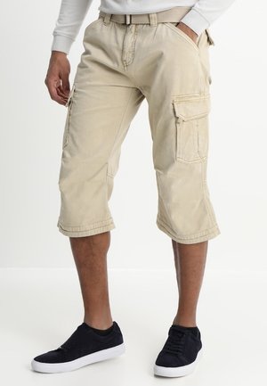 Mann trägt beige Cargo-Shorts mit Gürtel und schwarzen Sneakers, steht mit einer Hand in der Tasche vor weißem Hintergrund.