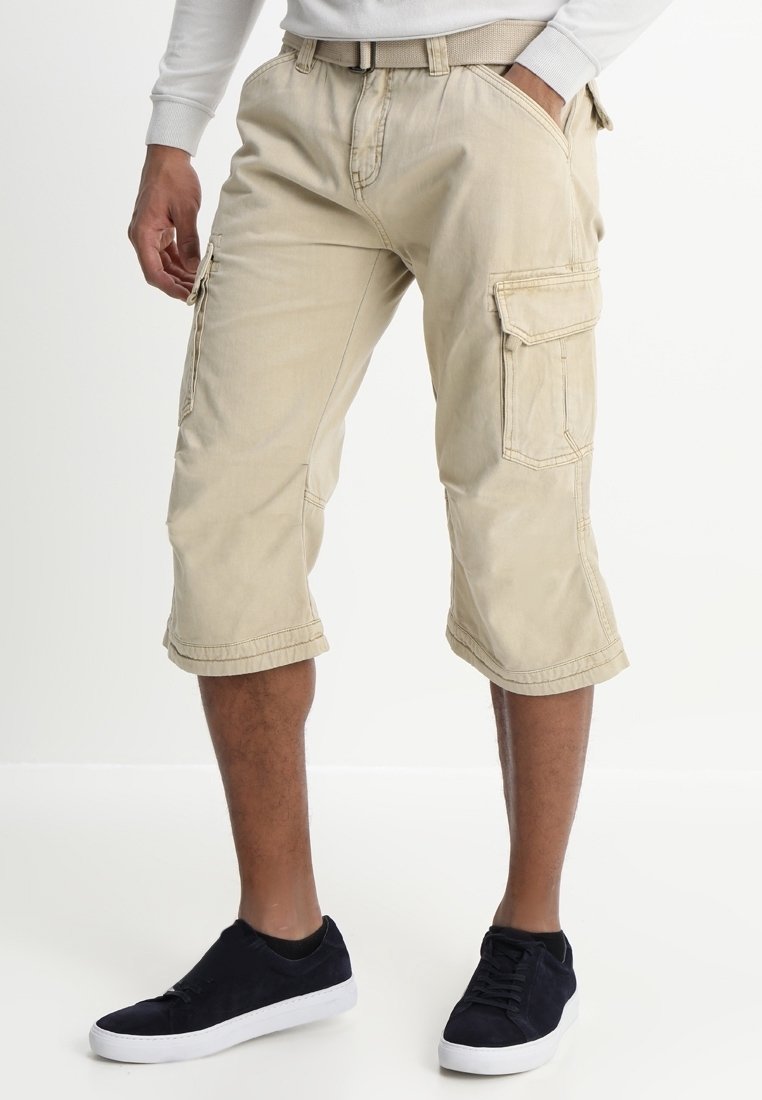 Homme portant un short cargo beige avec une ceinture et des baskets noires, debout une main dans la poche sur fond blanc.
